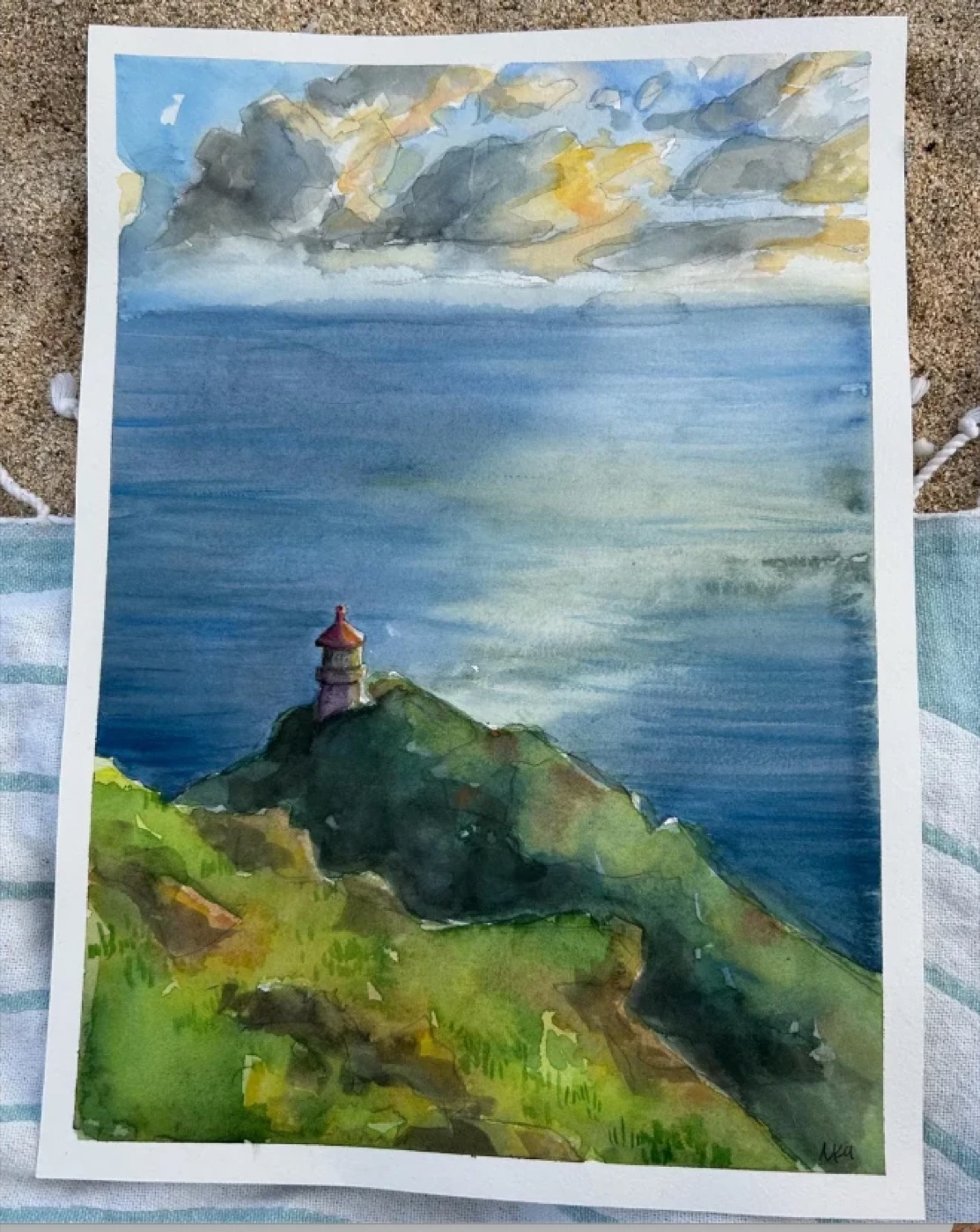 Watercolor of a lighthouse on a green cliff overlooking the ocean under a cloudy sky